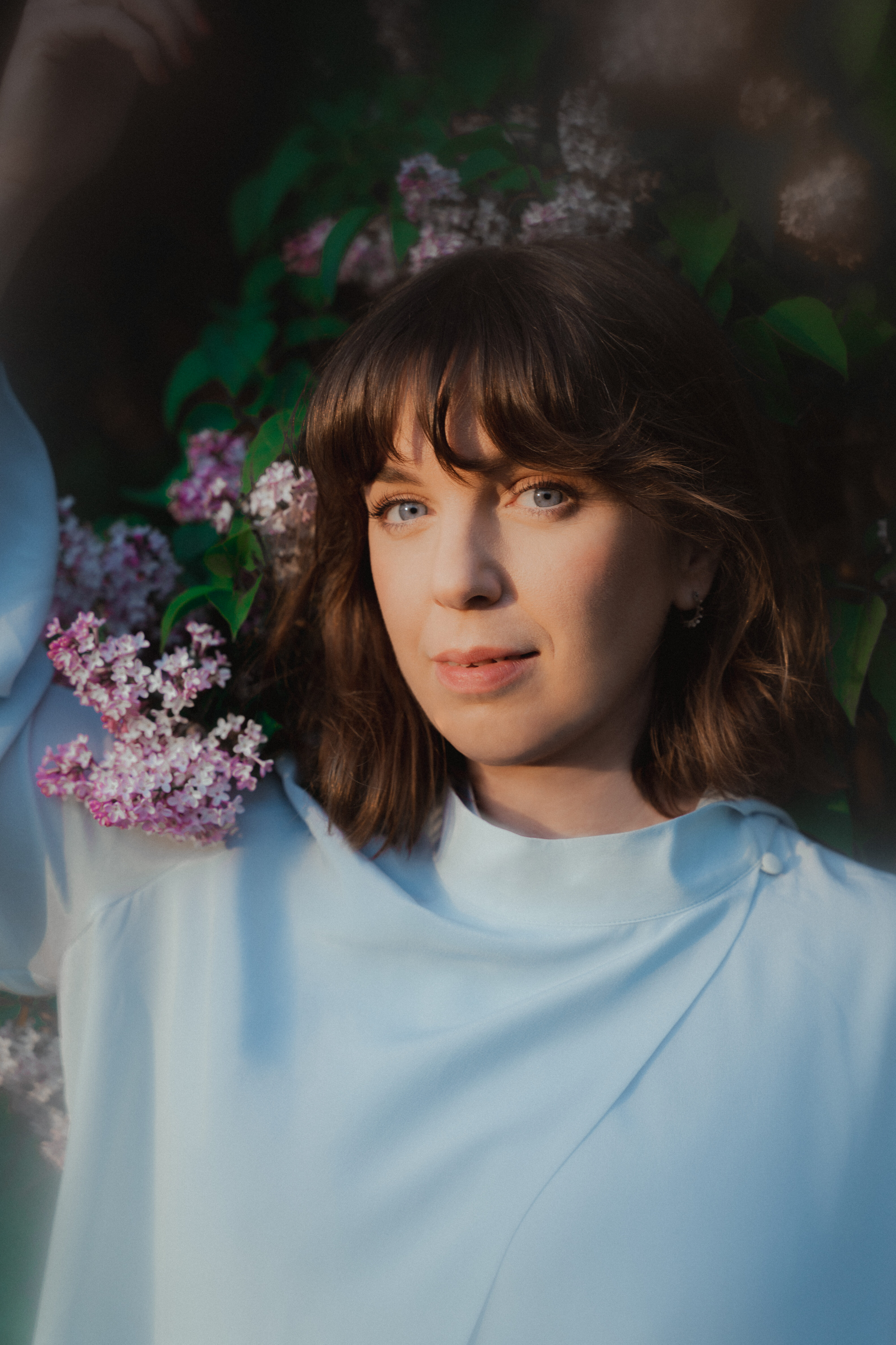 Woman with brown hair and blue eyes lying among pink lilac blossoms, wearing a light blue top; soft sunlight on her face.